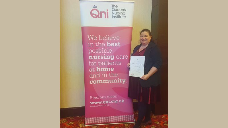 A woman stands in front of a banner with the logo of the Queen's Nursing Institute on it. She is holding a certificate and smiling.