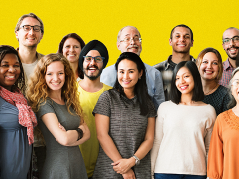 A group of men and women standing in a group smiling at the camera against a yellow background.