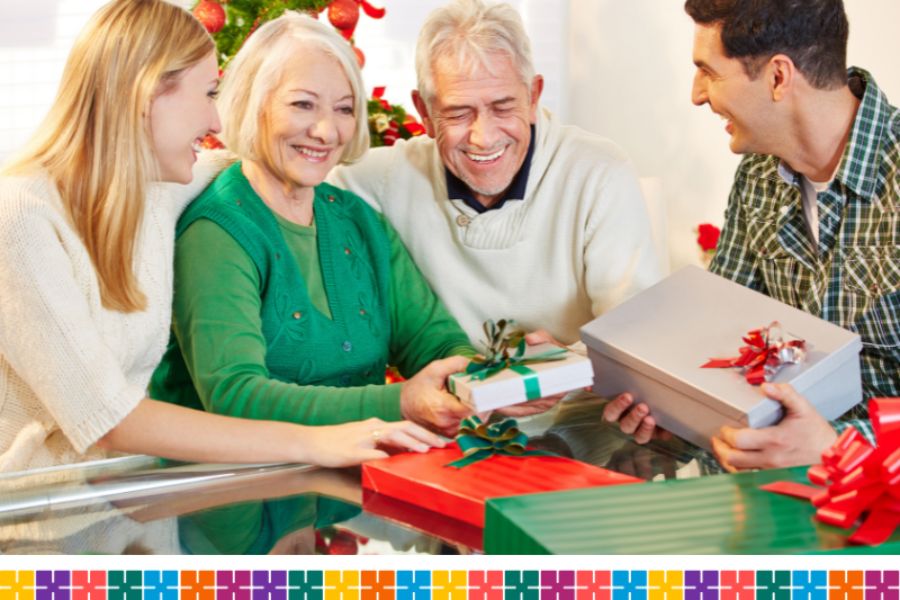 A young couple sitting either side of an older couple with a Christmas tree in the background, exchanging presents