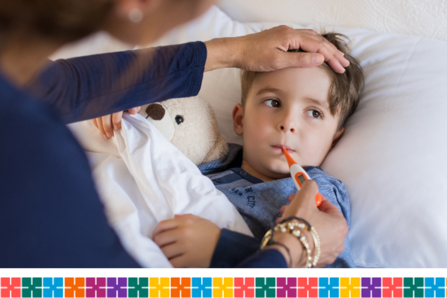 A child lying in bed cuddling a teddy bear, while their parent checks his temperature.