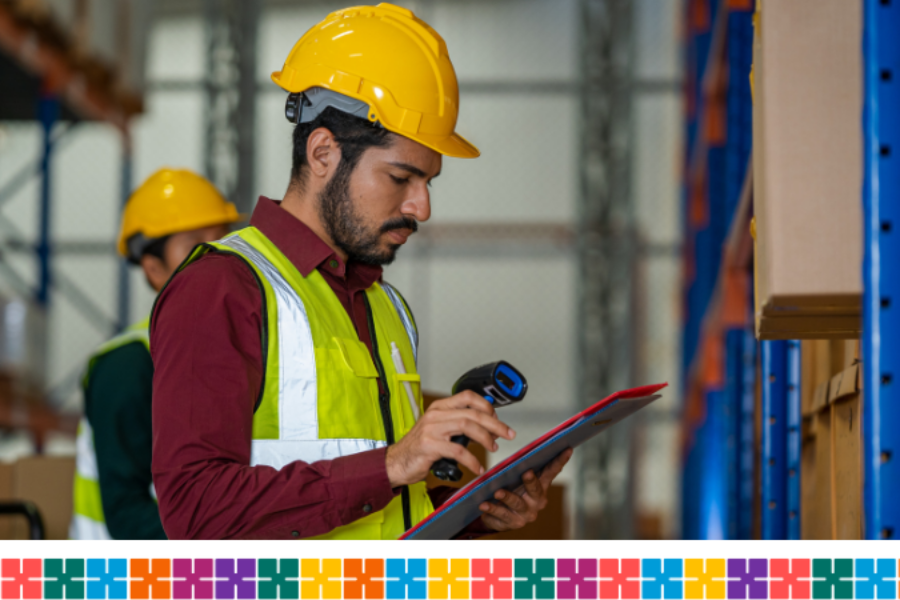 Male warehouse worker wearing a hard yellow hat and wearing a fluorescent jacket is looking down at a clipboard with a small scanning device in his right hand.