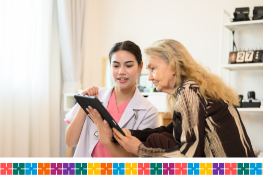 A younger woman with a stethoscope is showing information to an older woman. Both are seated in front of a bookcase and window.