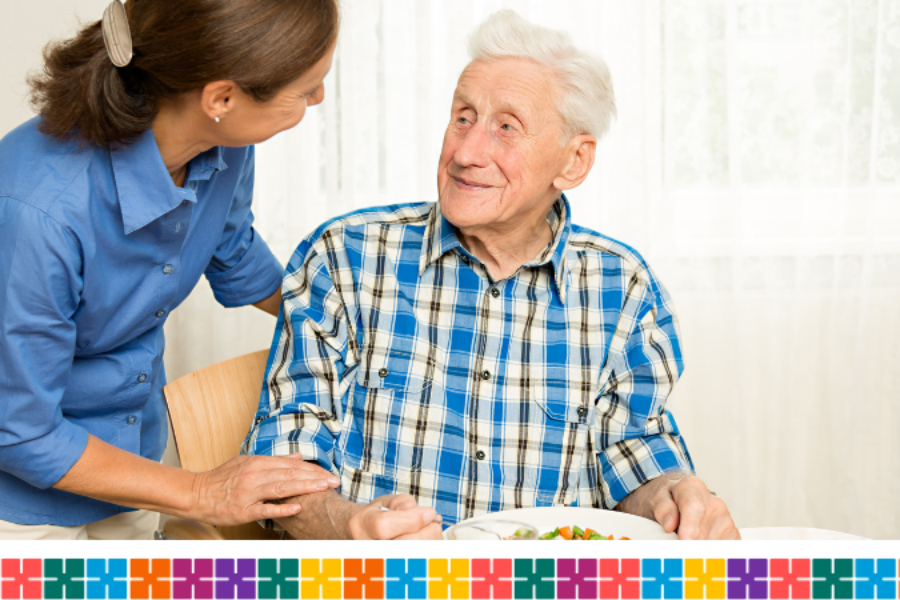 Elderly man sitting at a table with a plate of food and a woman stood next to him