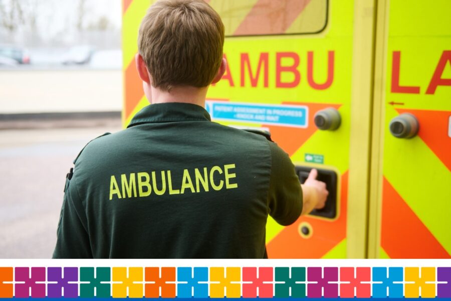 Back view of an ambulance crew member with the word ambulance on back of his shirt standing next to an ambulance