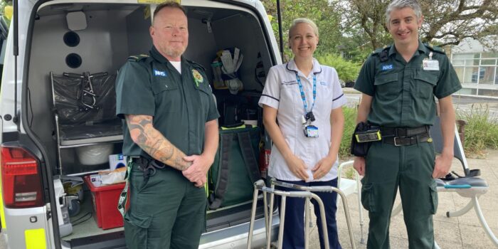 Three medical professionals - two men and one woman in the middle, stand smiling at the camera in front of a van which is open and contains medical equipment.