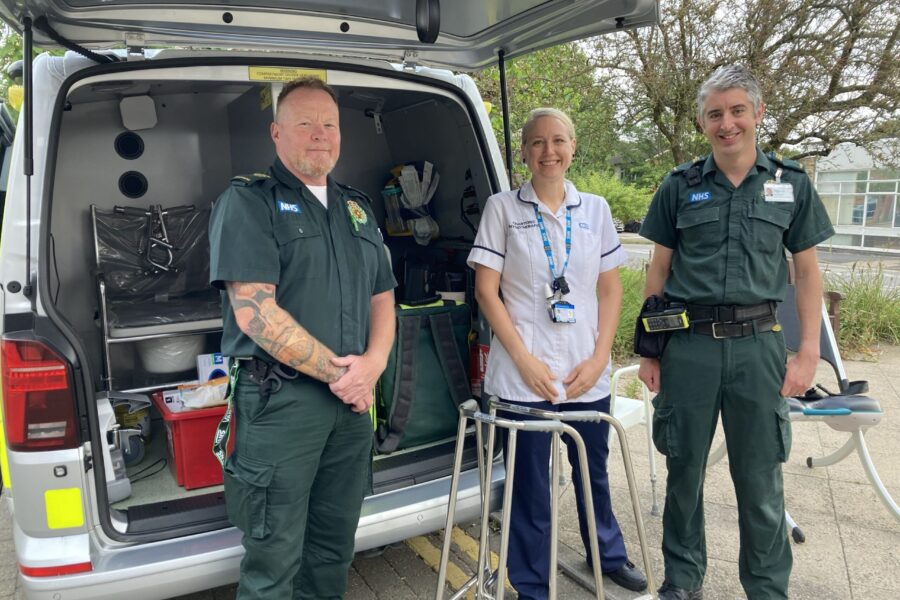 Three medical professionals - two men and one woman in the middle, stand smiling at the camera in front of a van which is open and contains medical equipment.