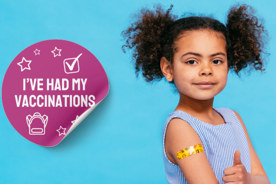 Young girl looking at the camera with her thumb up. She has a yellow sticker on her arm. Next to her is a big purple sticker that says "I've had my vaccinations".