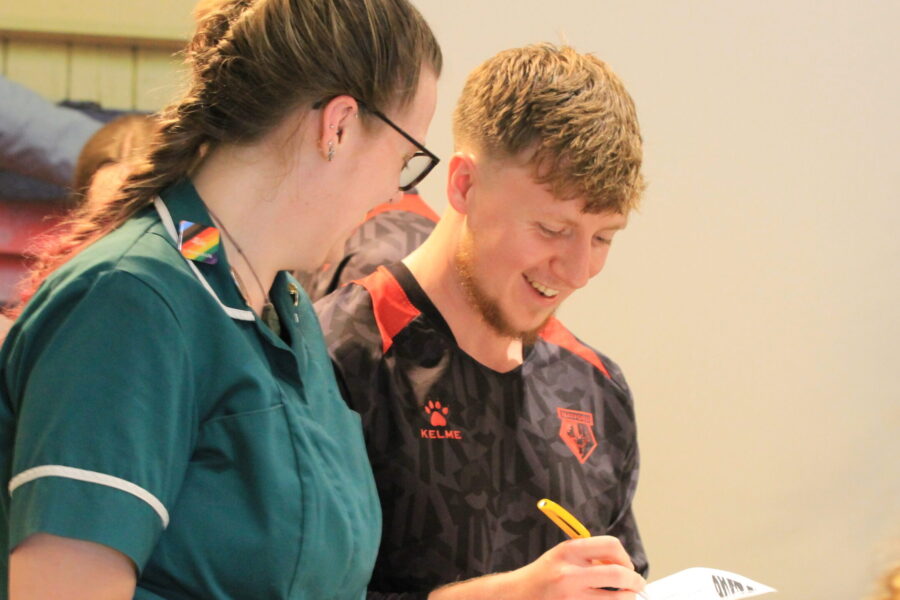 Nurse wearing a green uniform speaking to a member of Watford Football Club who is wearing the club's shirt whilst filling in a clip board.